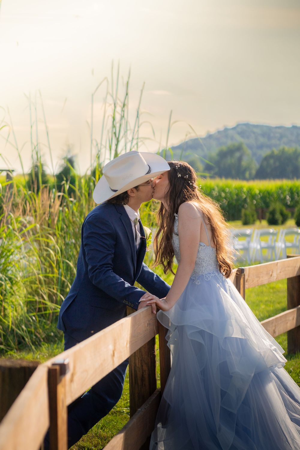 A romantic couple in wedding attire share intimate moments along a rustic wooden fence with cornfields and mountains in view.