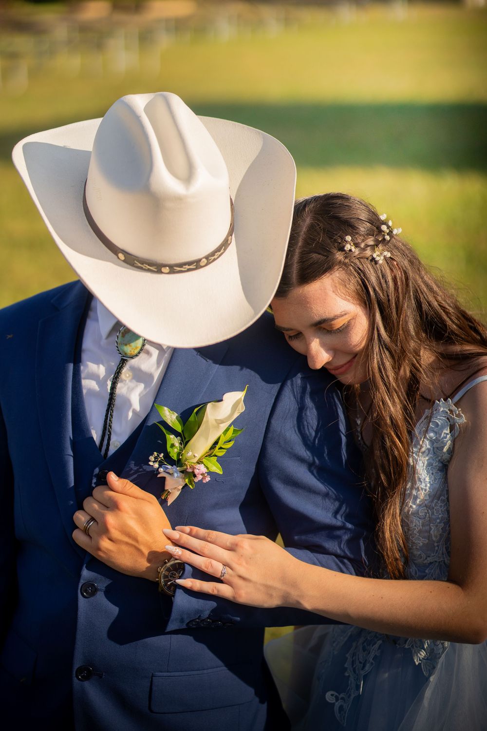 A romantic country wedding moment showing a white cowboy hat, navy suit, and floral boutonniere in warm sunlight.