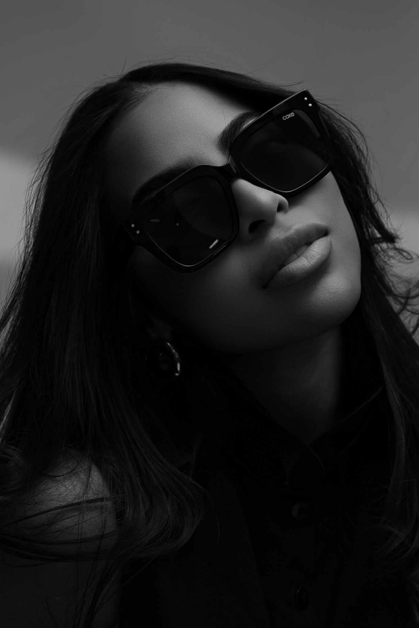 A close-up black and white portrait of a woman wearing large square sunglasses with long dark hair.