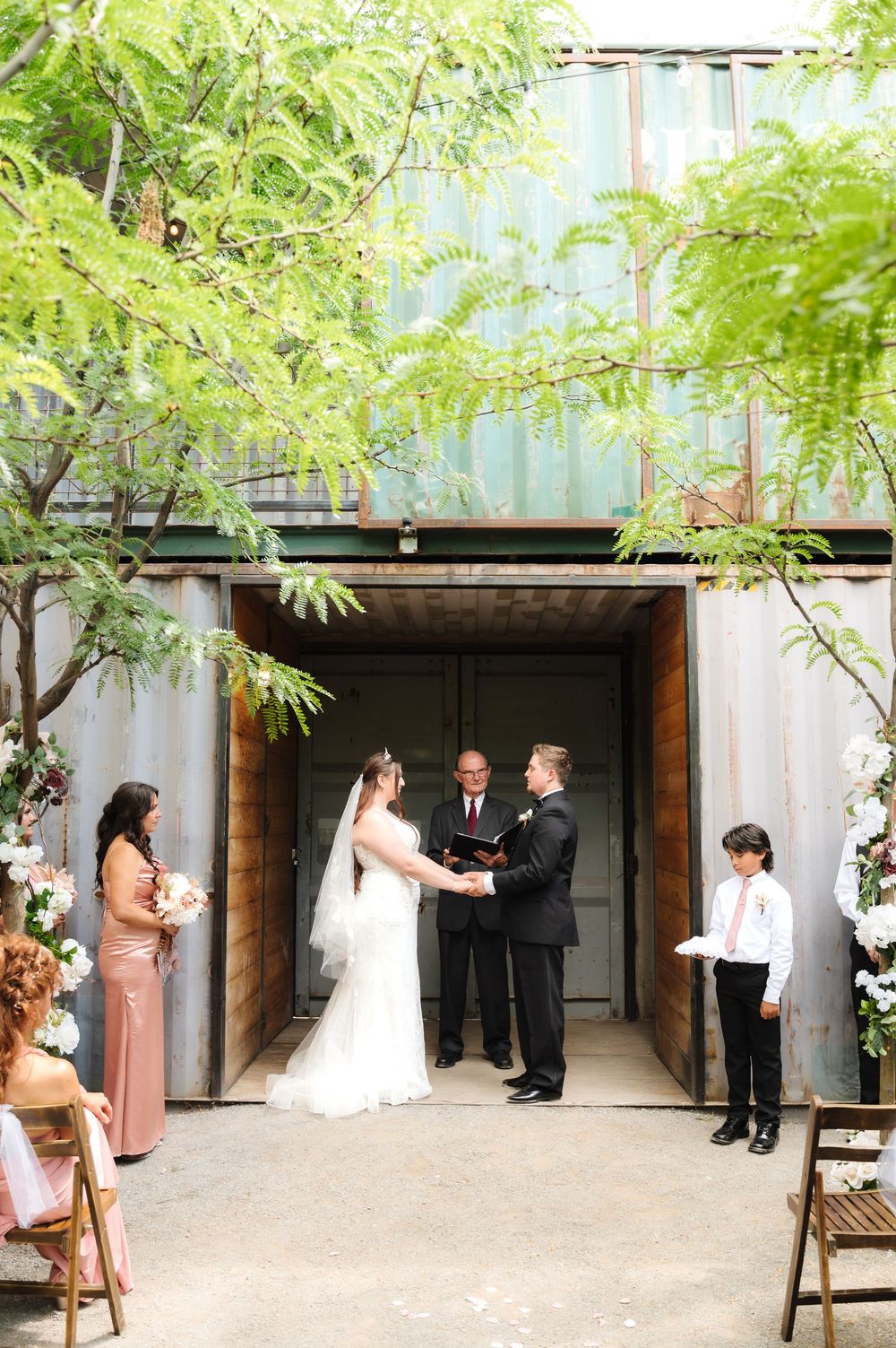 A wedding ceremony unfolds in a courtyard with green trees overhead and brick walls creating an intimate atmosphere.