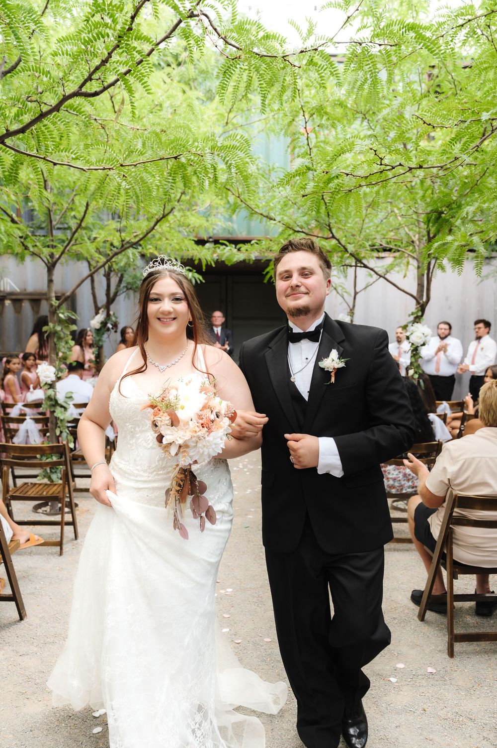 A couple dances joyfully under green trees at an outdoor wedding reception venue.