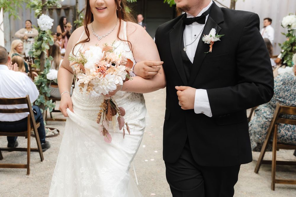 A couple walks arm in arm at their wedding ceremony, with the bride holding a peach and white floral bouquet.