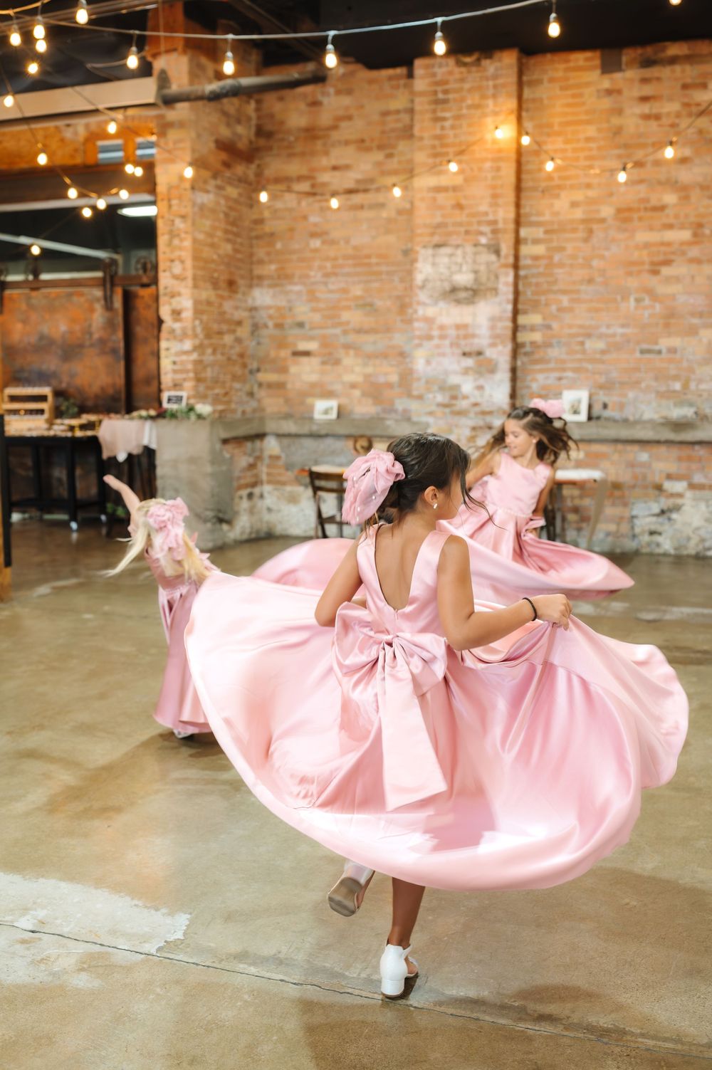 A person in a flowing pink dress twirls gracefully in an industrial brick-walled space with string lights overhead.