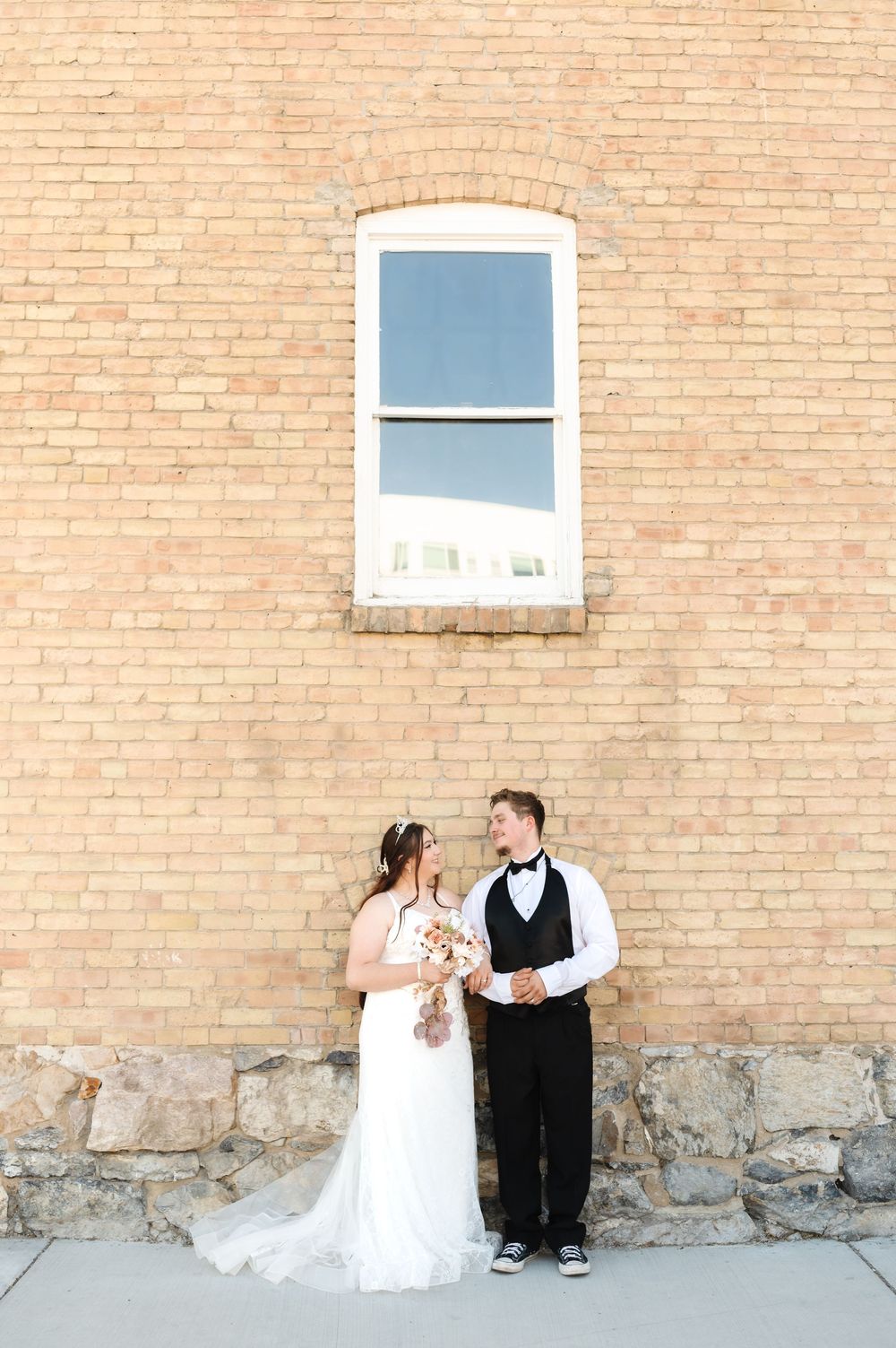 A wedding couple poses against a rustic brick wall with small windows for their portrait session.