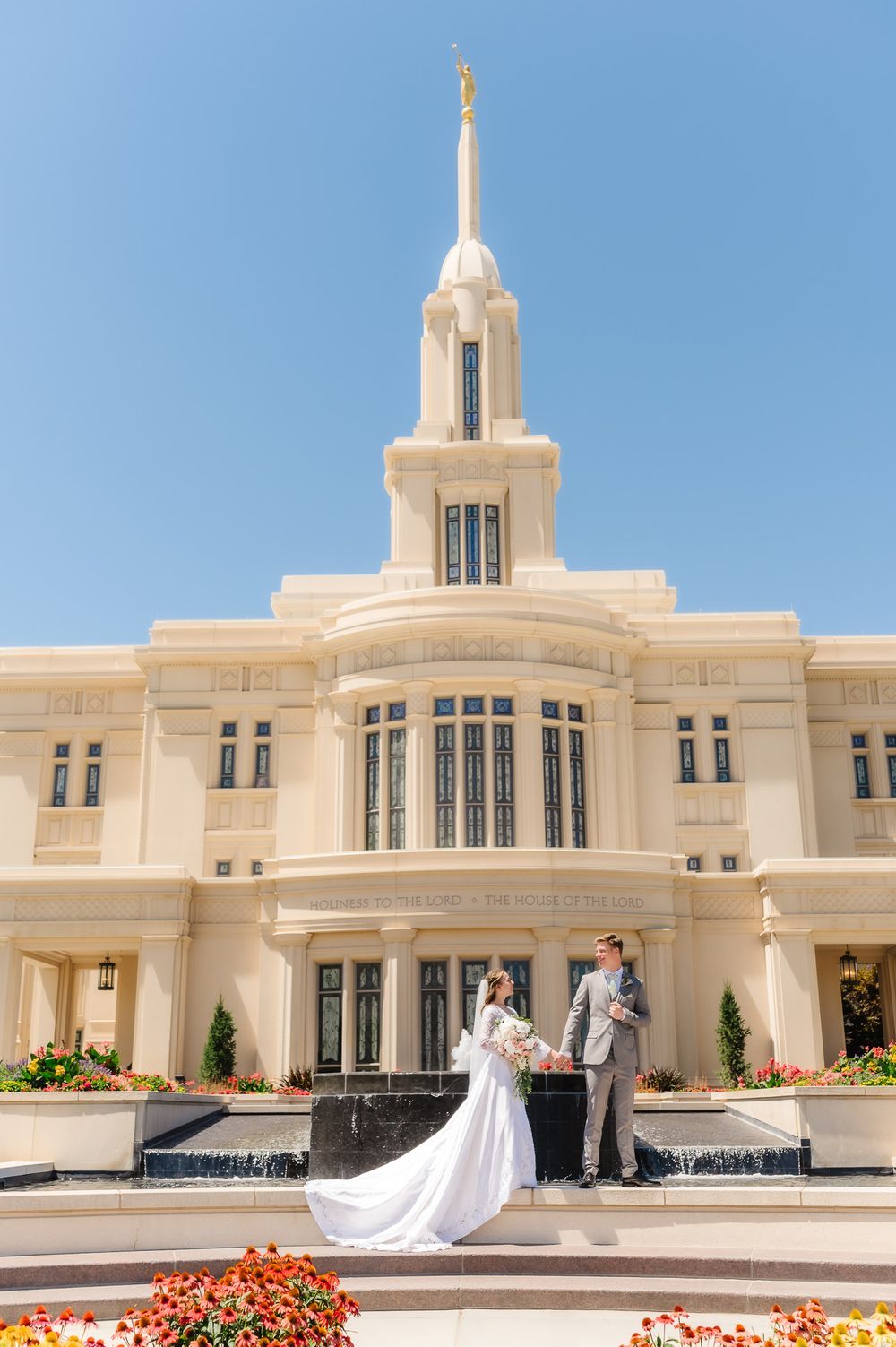 A wedding couple poses on the steps of a grand white temple with ornate architecture and colorful flower beds.