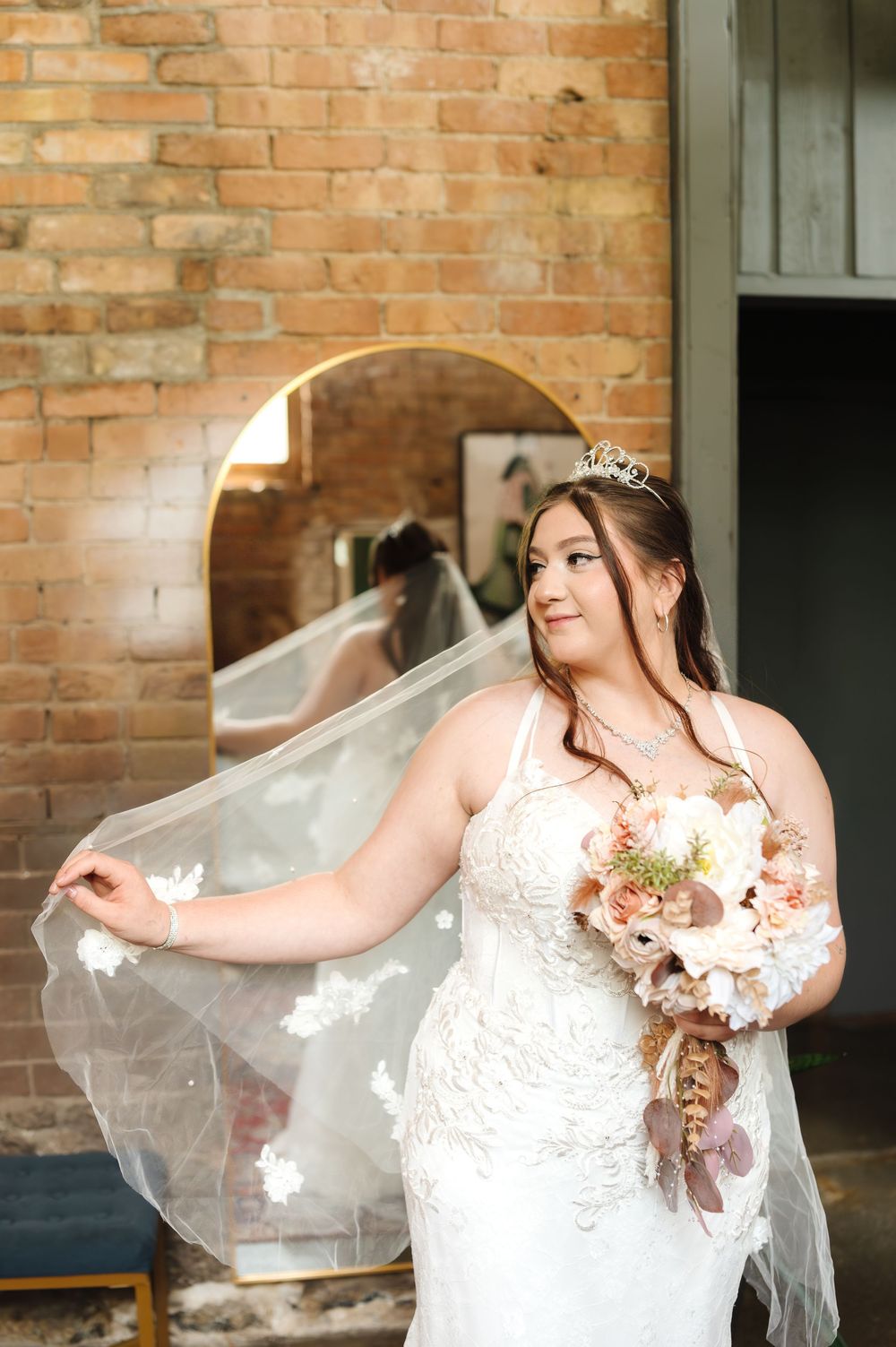 A bride in a white lace gown with a tiara holds her flowing veil against a rustic brick wall backdrop.