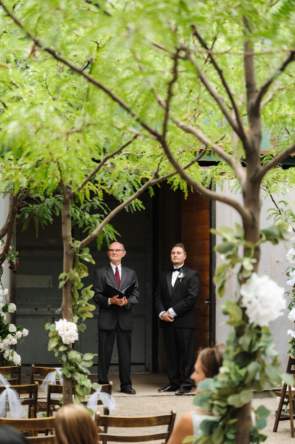An outdoor wedding ceremony setup with wooden chairs beneath leafy green trees.