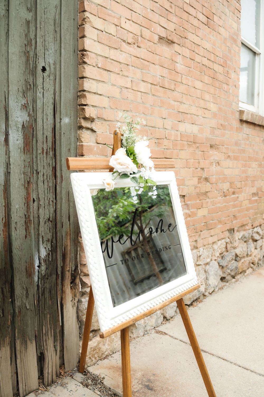 A white framed mirror sign with floral decor sits on a wooden easel against an exposed brick wall.