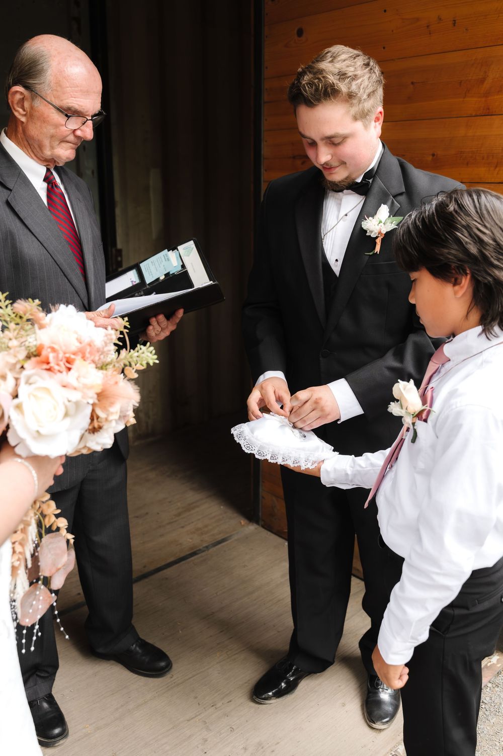 Wedding party members sit on wooden benches holding floral bouquets during an outdoor ceremony.