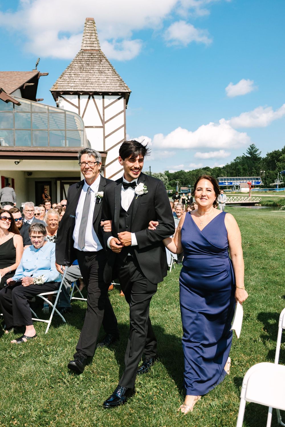 Wedding party members walking on grass lawn with white tower building and blue sky in background.