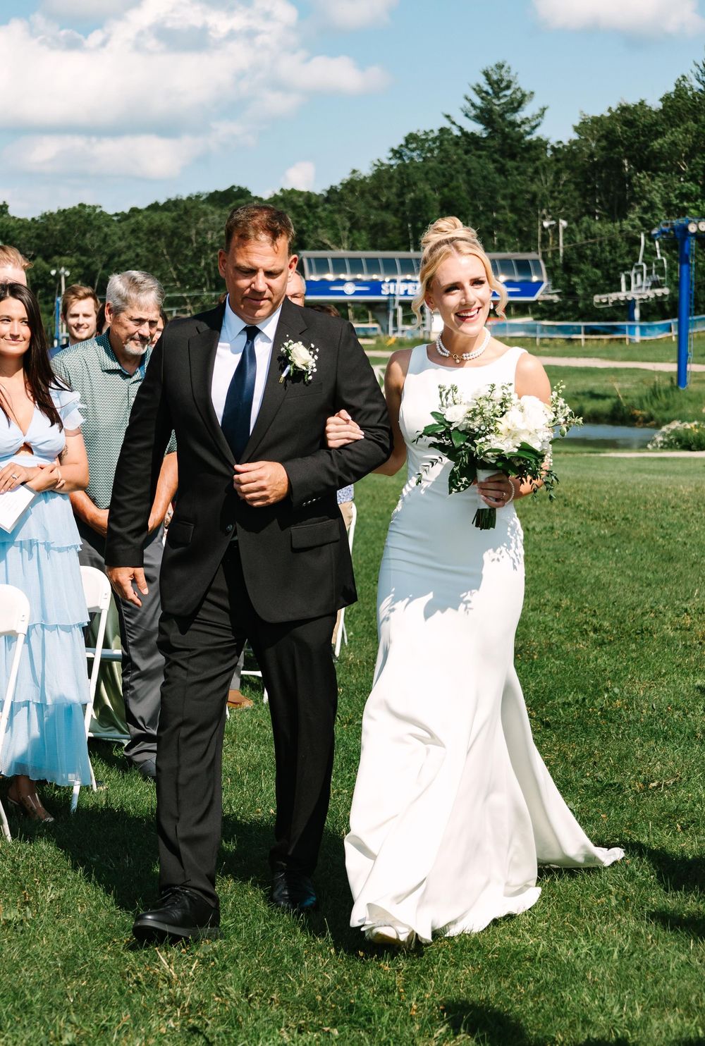 Outdoor wedding ceremony procession on green lawn with lake and trees in background.