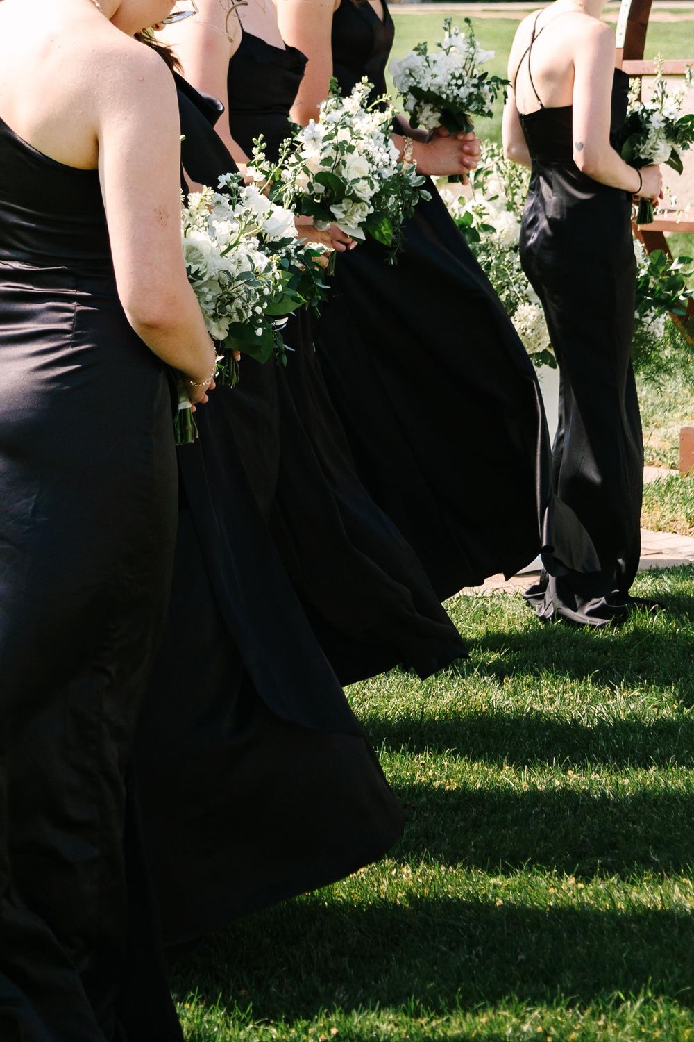 Row of bridesmaids in black dresses holding white bouquets stand on grass during outdoor wedding ceremony.