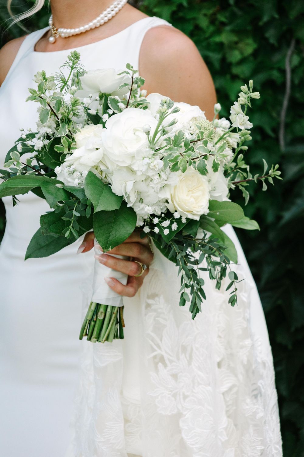 A white bridal bouquet featuring roses and delicate greenery held against an elegant white wedding dress.
