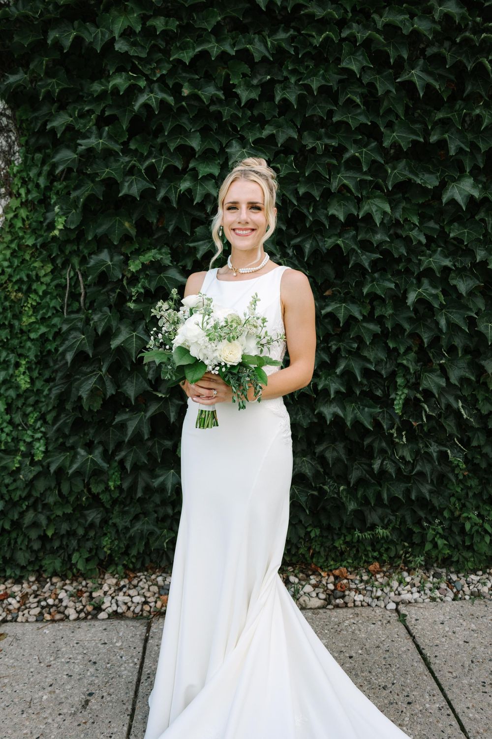 A bride in a sleeveless white wedding dress poses with a green and white floral bouquet against an ivy-covered wall.