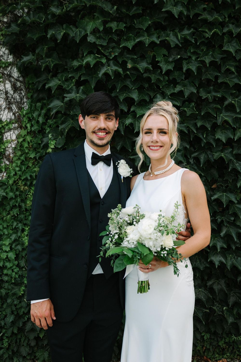 Wedding couple poses against ivy-covered wall in formal attire with white floral bouquet.