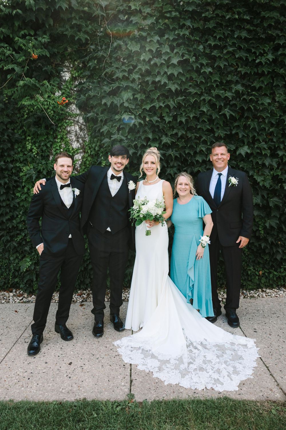 Wedding party poses together in front of green ivy wall wearing formal black suits and turquoise bridesmaid dress.