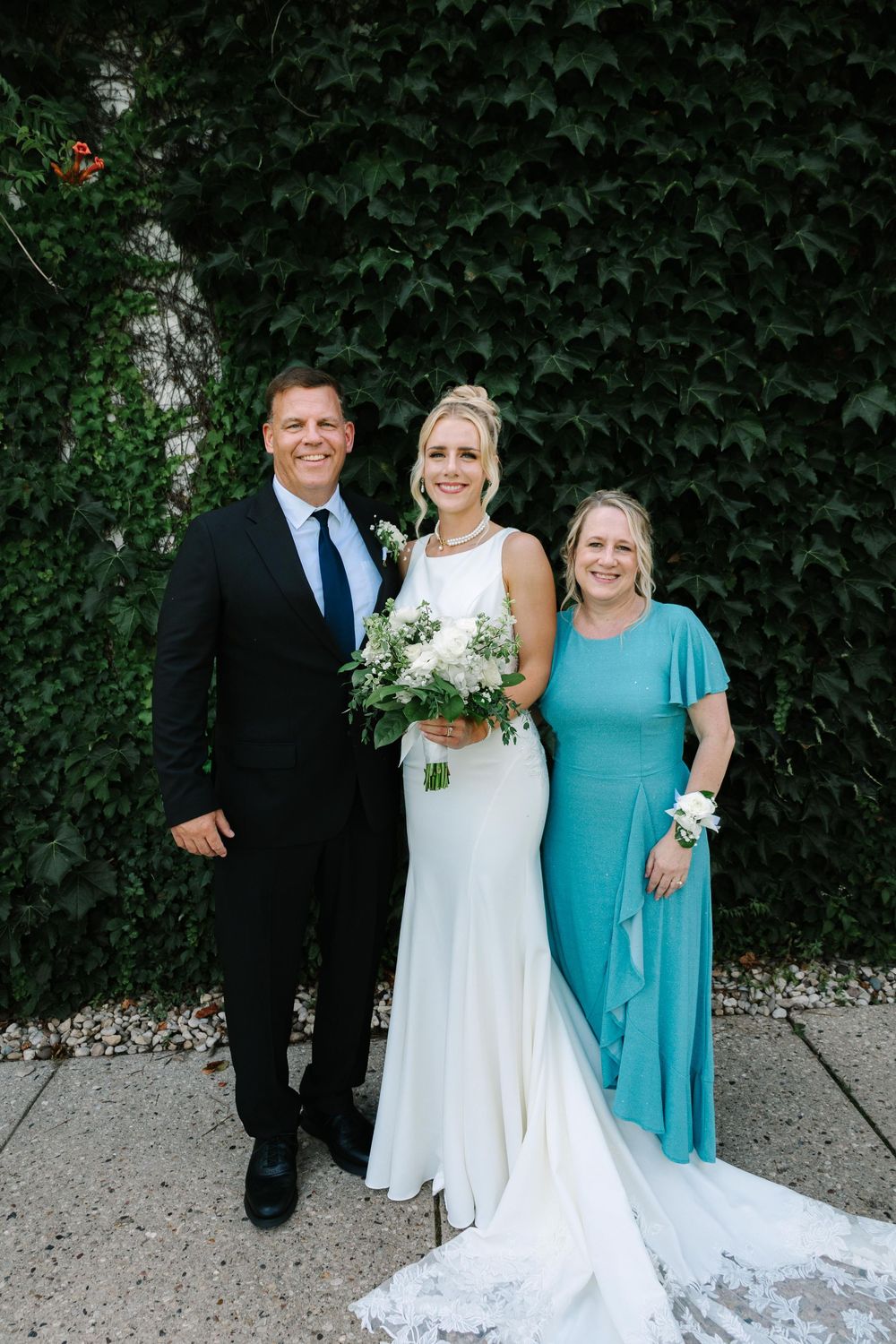 Wedding group photo with bride in white gown and attendant in turquoise dress against green hedge.