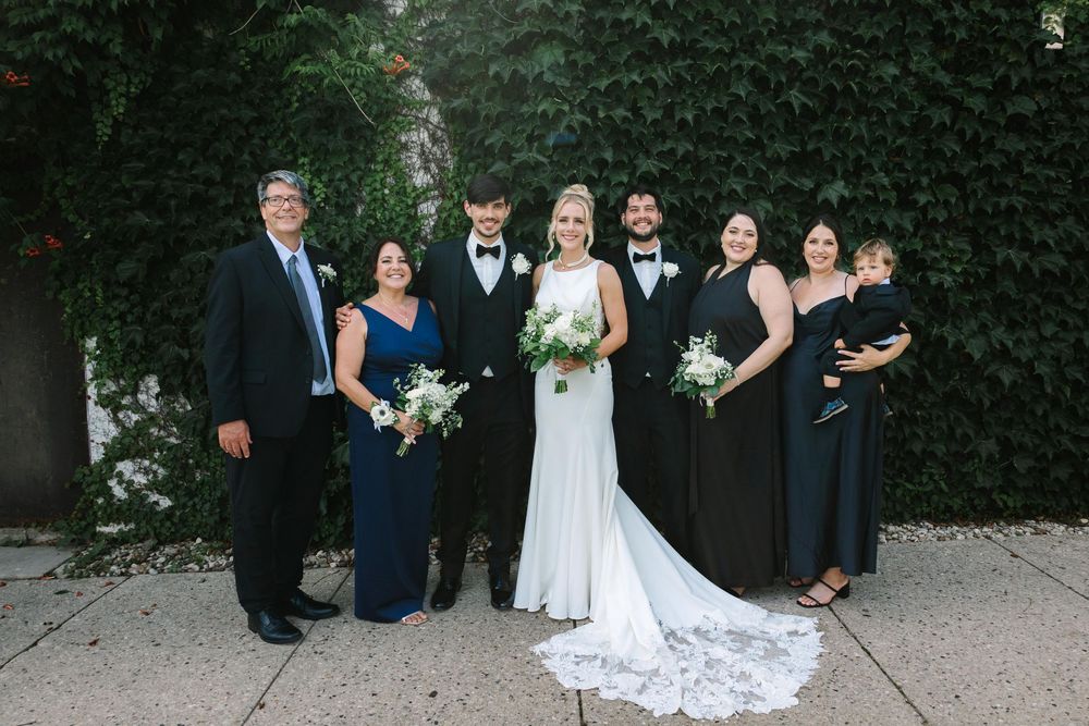 Wedding party poses together in formal attire on garden pathway during outdoor celebration.