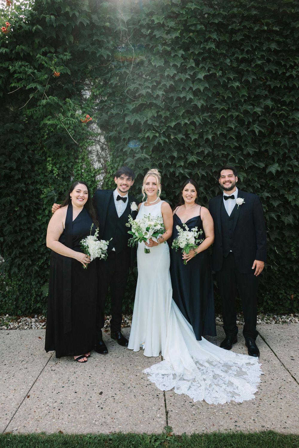Wedding party members stand together against lush green foliage backdrop in formal attire.