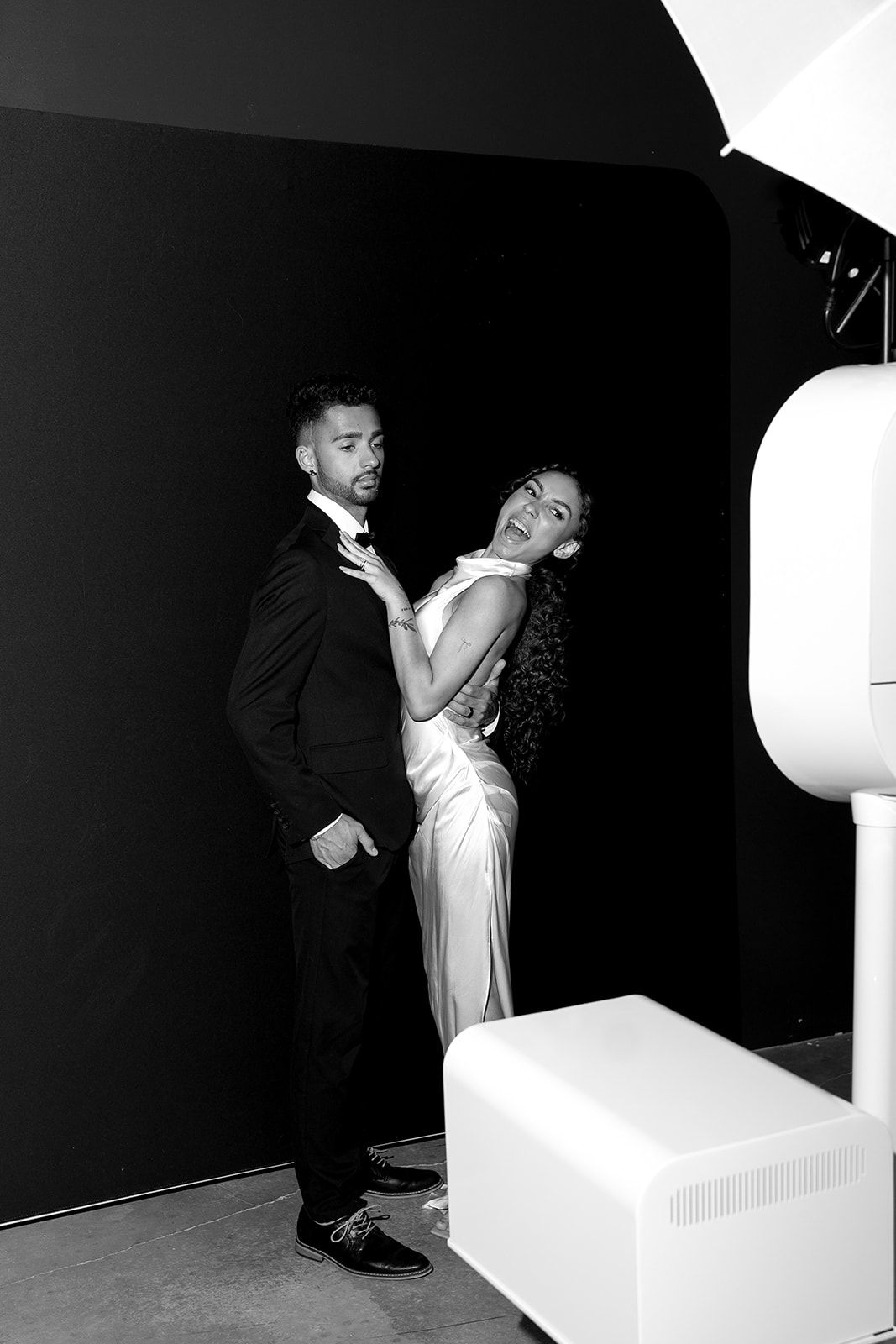 A couple poses in black and white, with the woman in a white dress leaning back against her partner in a tuxedo, in front of a photo booth.