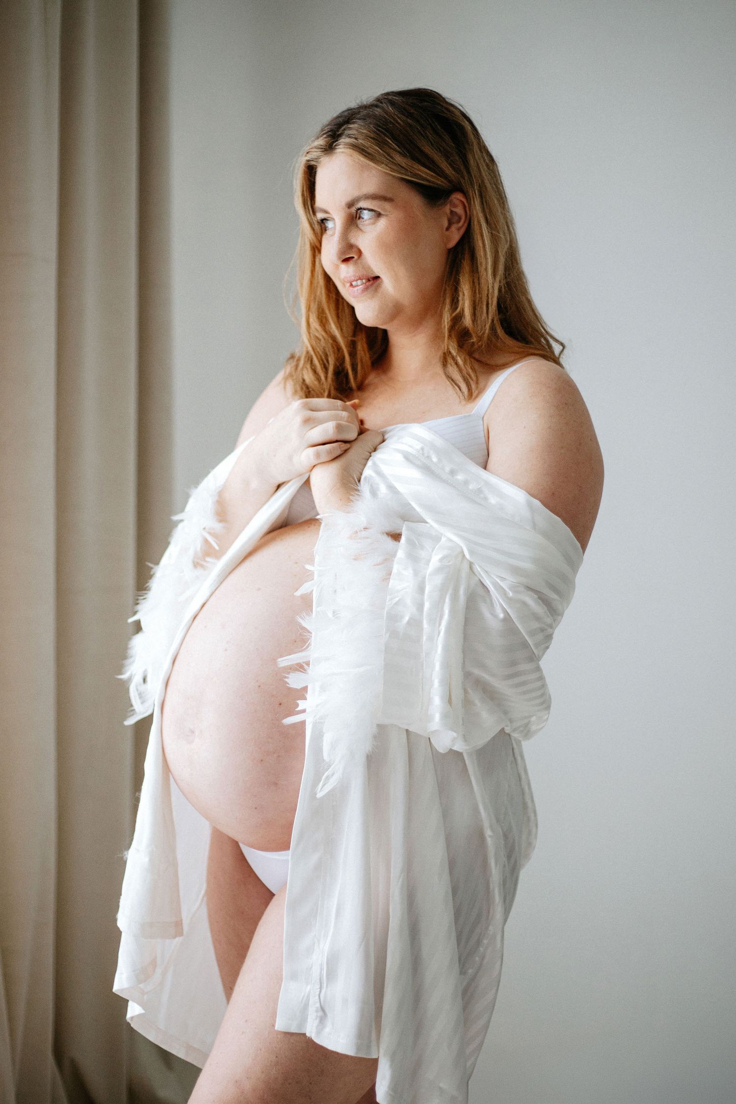 Maternity photo of an expectant mother in white feathered robe standing by window in soft natural light.
