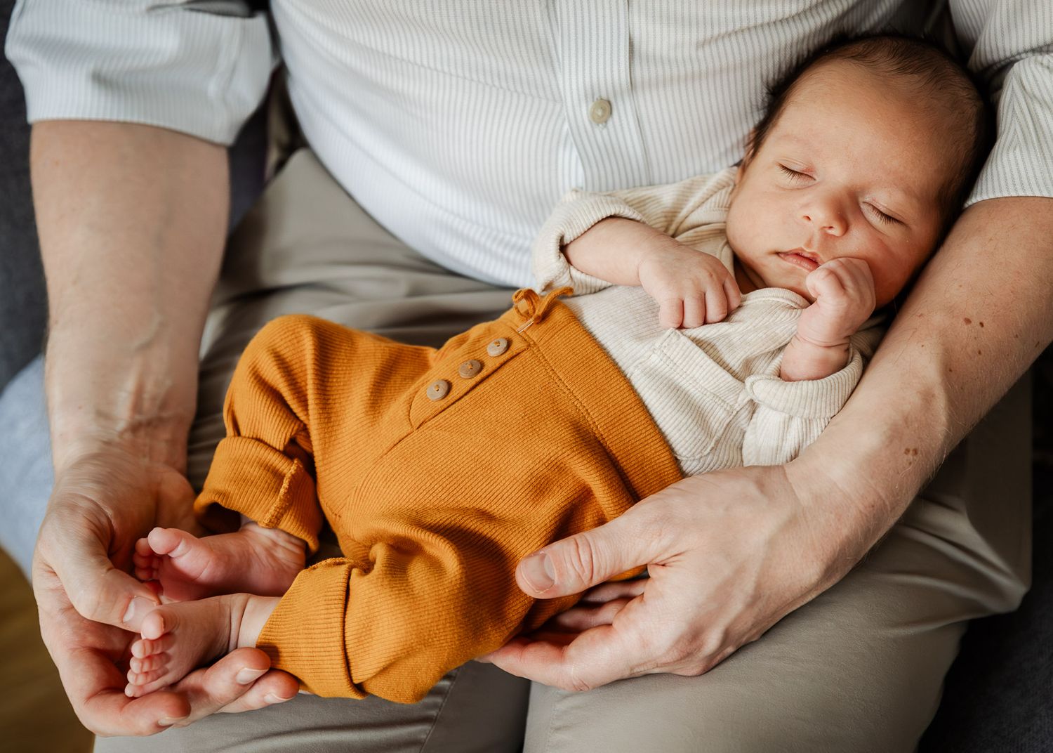 A newborn baby sleeps in his fathers lap during an in-home motherhood photoshoot in Bristol with Little Bodhi Photography