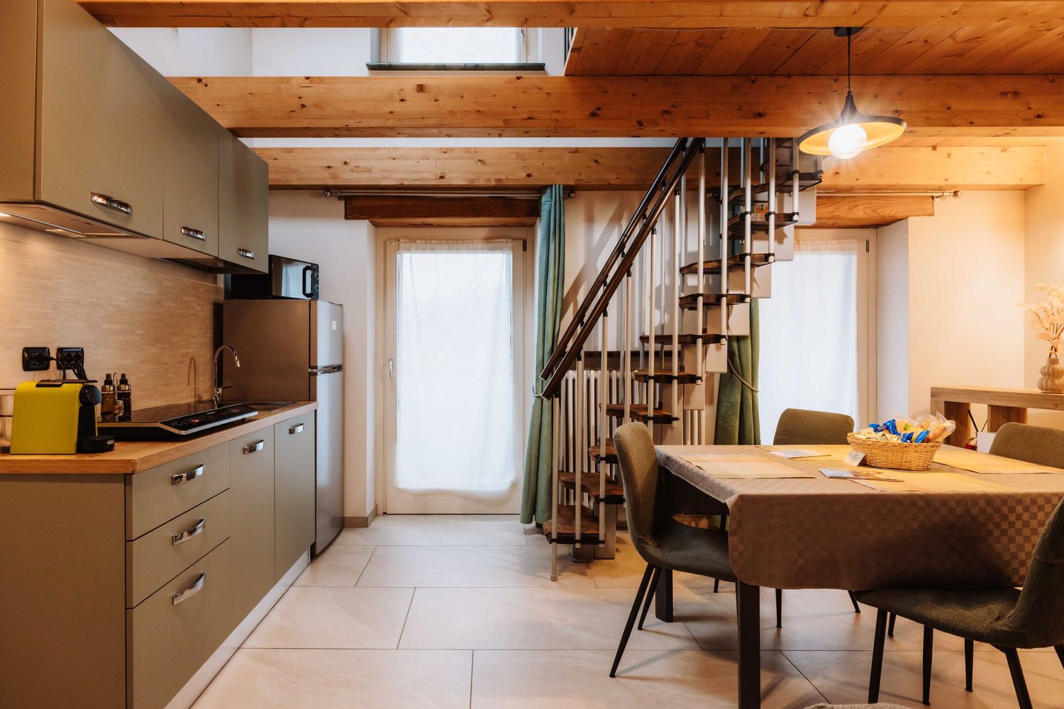 Modern kitchen and dining area with wooden beams, gray cabinets, and a spiral staircase in a cozy apartment space.