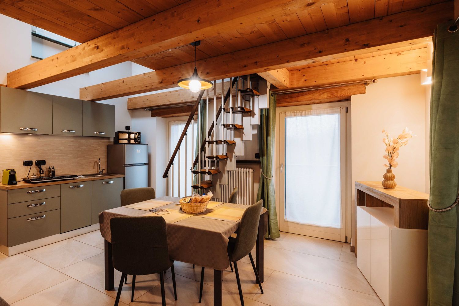 Modern kitchen and dining area with wooden beams, spiral staircase, and gray cabinets in a cozy interior space.