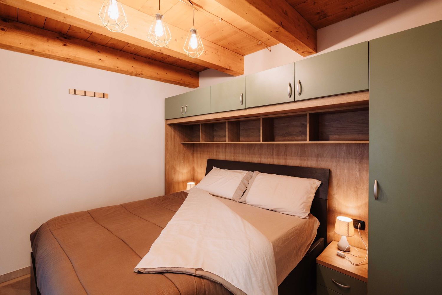Modern bedroom with built-in storage headboard, exposed wooden beams, and sage green cabinets above the bed.
