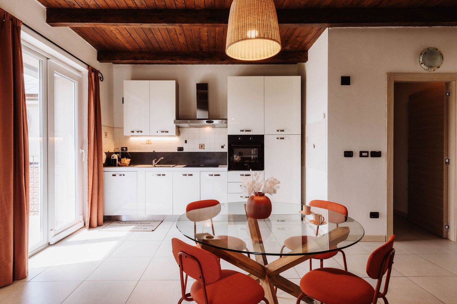 Contemporary white kitchen with glass dining table, orange chairs, and wooden beam ceiling in bright natural light.