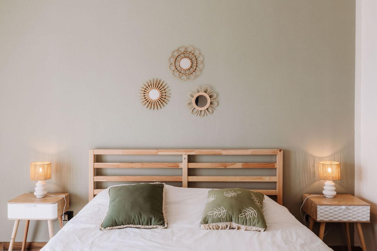 Modern bedroom interior with wooden headboard, green pillows, white side tables, and decorative rattan wall art above.