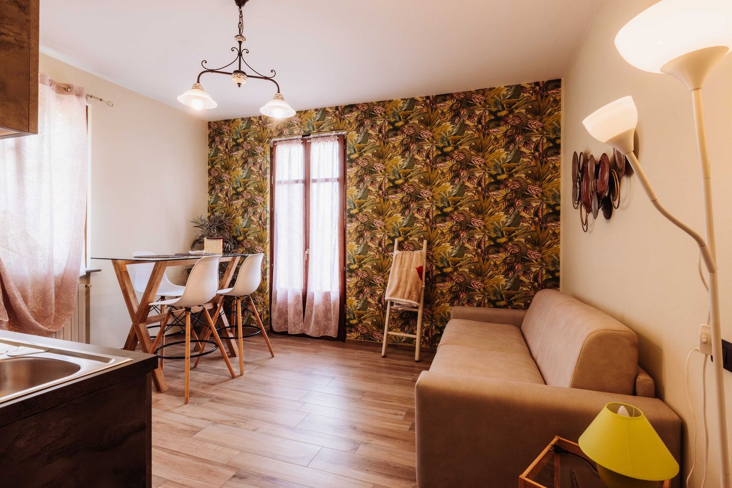 A cozy living room with floral wallpaper, hardwood floors, brown sofa, and tall windows with natural light.