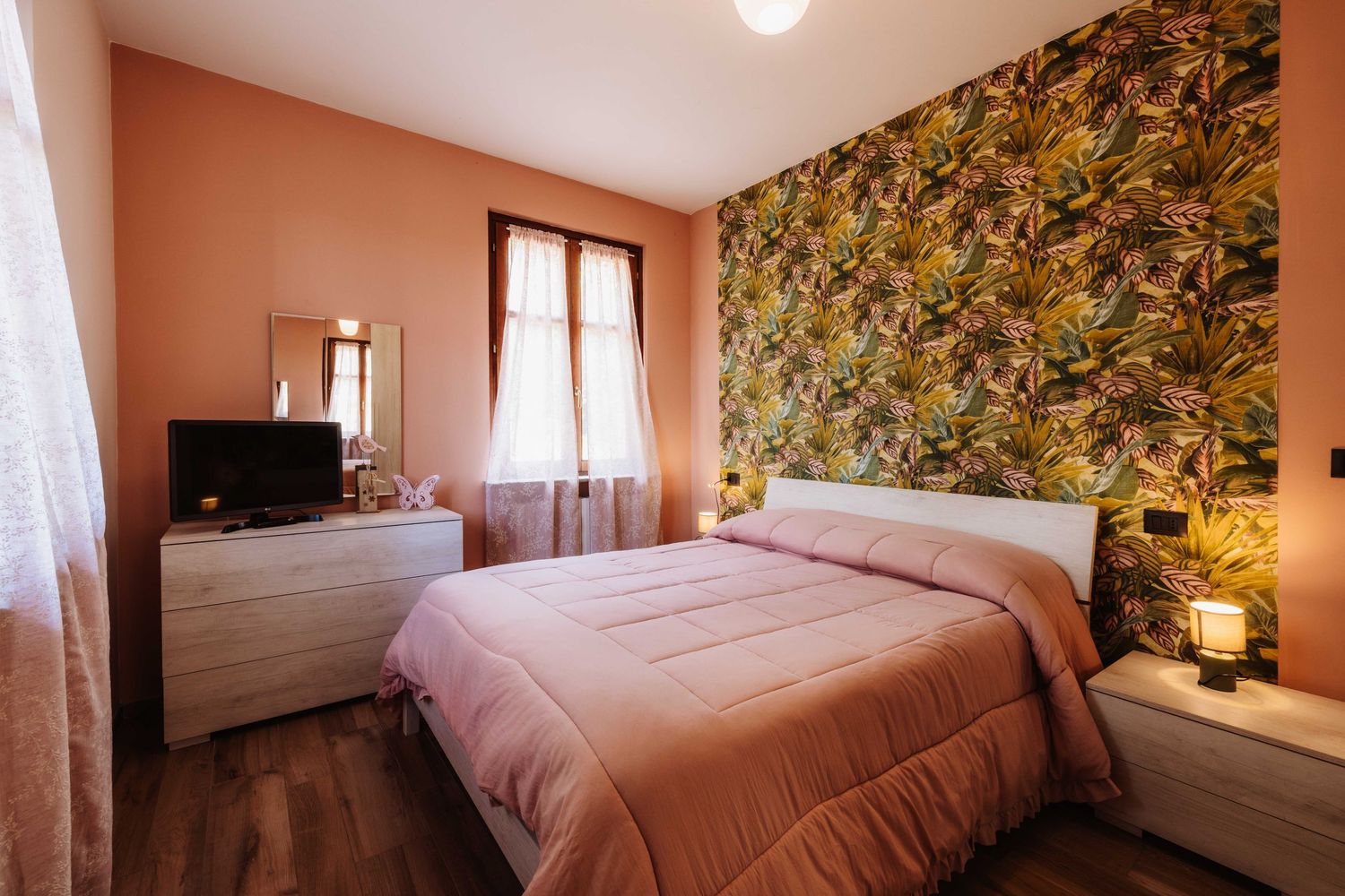 Cozy bedroom interior with floral wallpaper, pink bedding, and natural light streaming through sheer curtains.