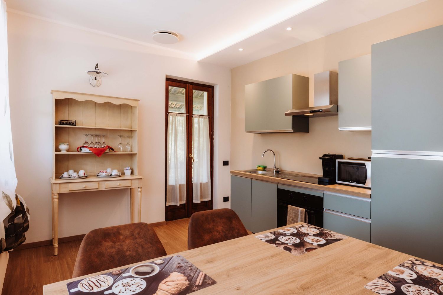 Modern apartment kitchen with wooden dining table, built-in shelves, and stainless steel appliances in neutral colors.