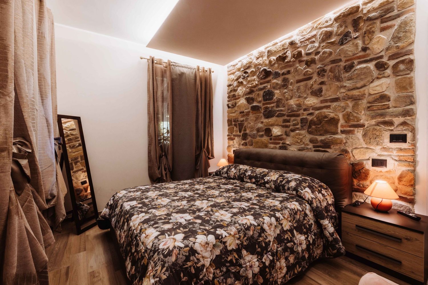 Rustic stone wall bedroom with modern lighting features a floral patterned bedspread and soft ambient glow.