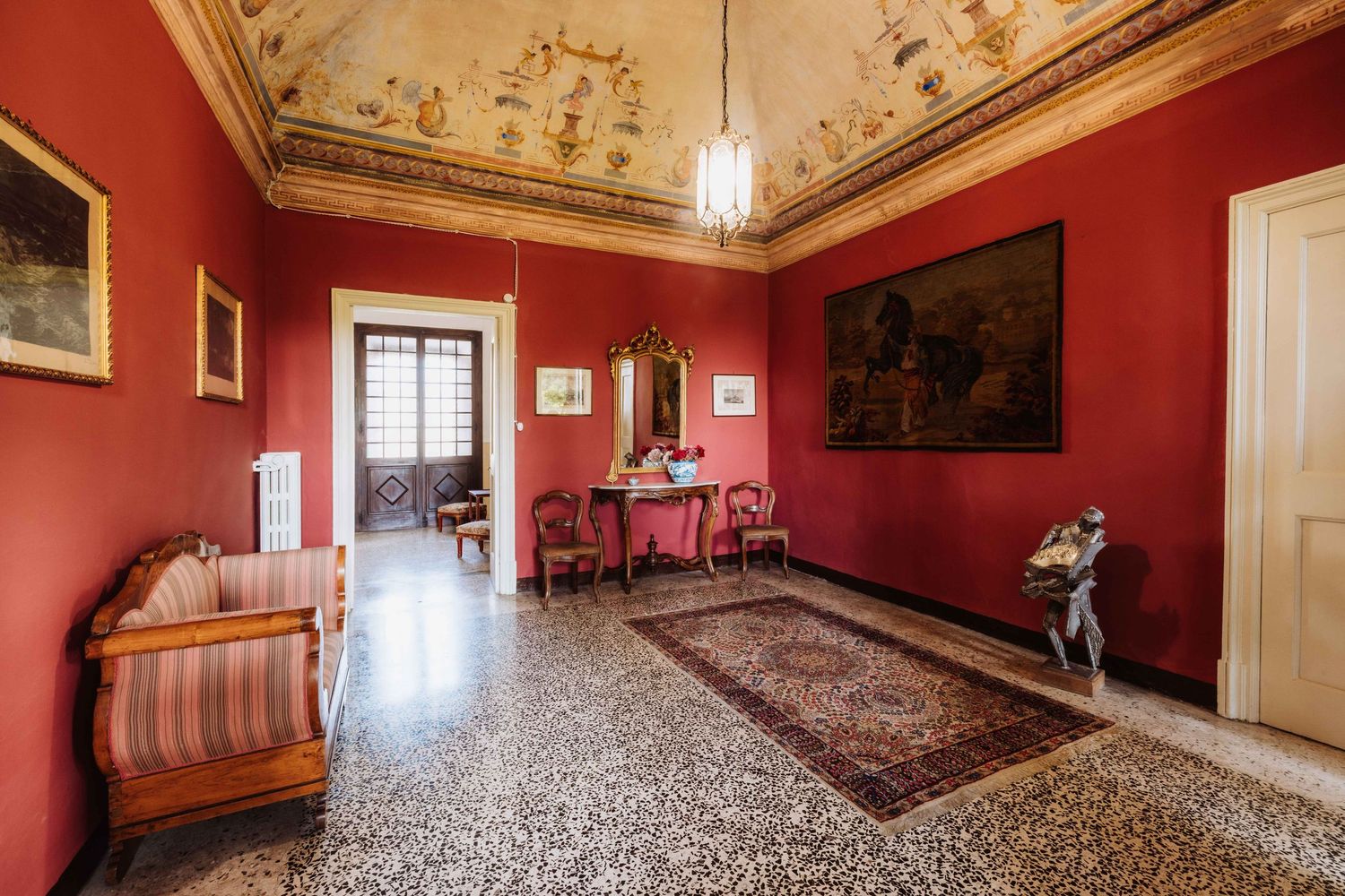 Elegant red-walled hallway with ornate ceiling, antique furniture, and traditional terrazzo flooring in a historic mansion.