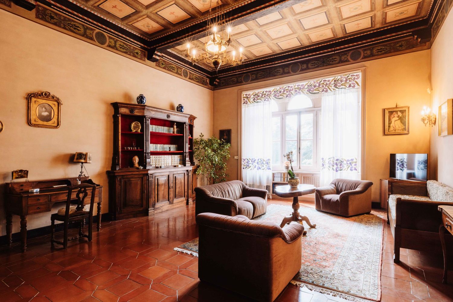 Elegant Victorian living room with ornate coffered ceiling, dark wood furniture, and large windows letting in natural light.