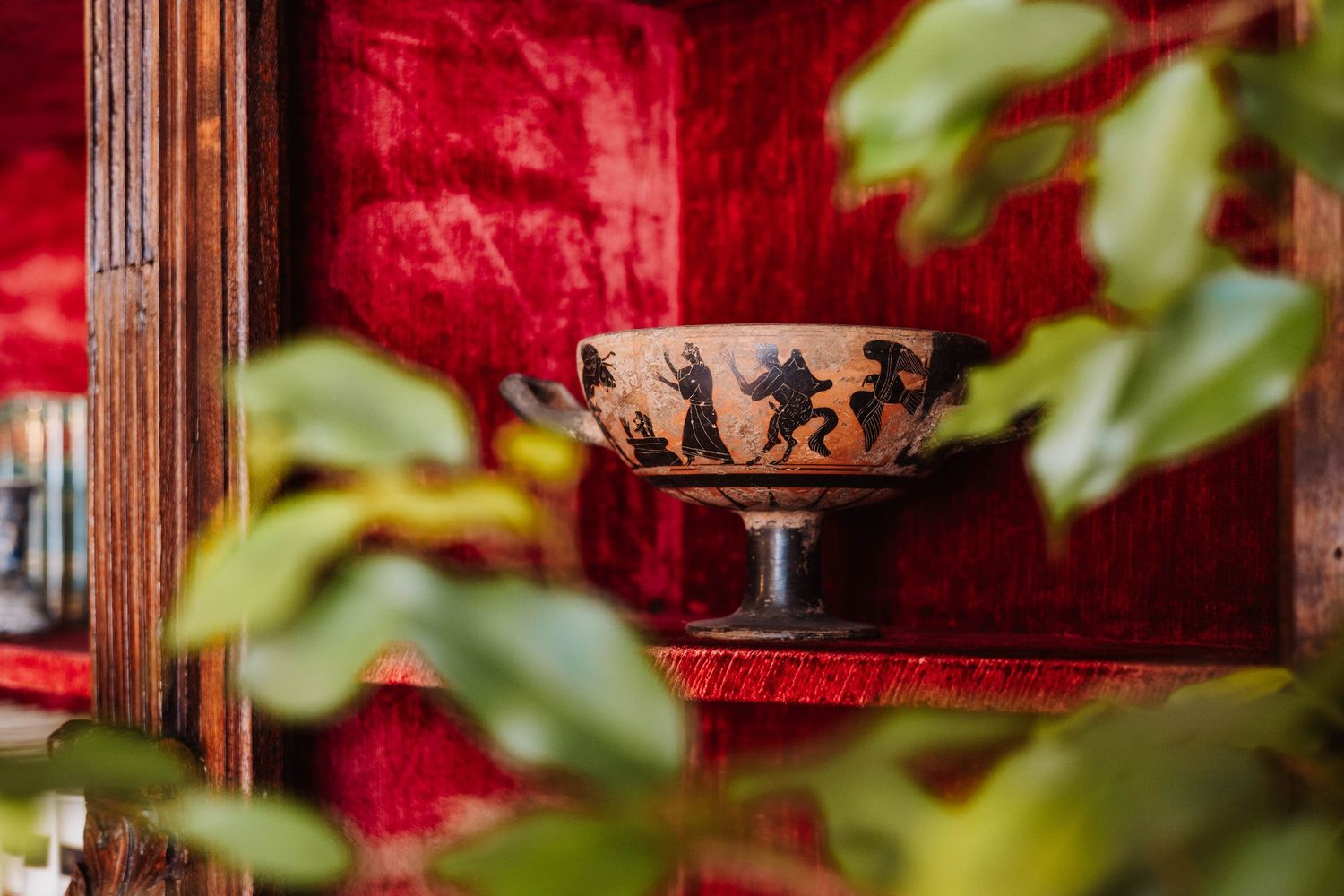 Ancient Greek style pottery bowl with black figures displayed against a red background with green leaves in foreground.