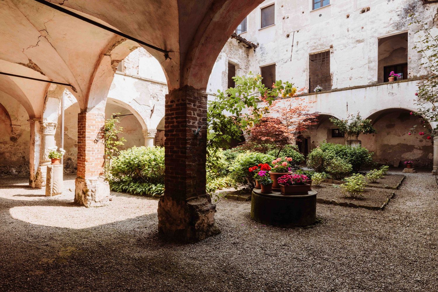 Ancient stone courtyard with arched colonnade and blooming flowers in terracotta pots under Mediterranean sunlight.