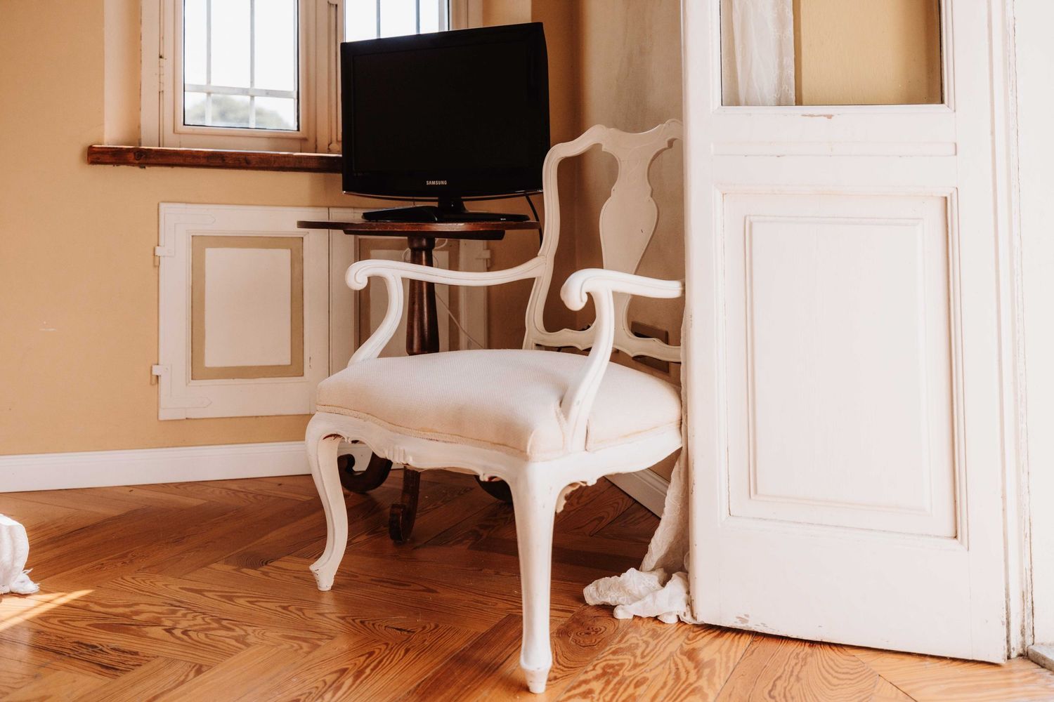 Elegant white vintage chair sits beside a small TV stand on hardwood floors in a room with peach-colored walls.