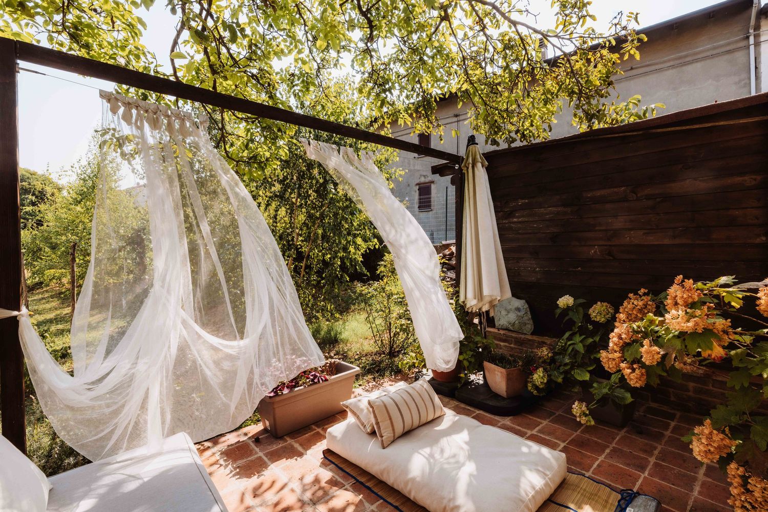 White sheer curtains blow in the breeze on a rustic wooden deck surrounded by lush greenery and orange flowers.