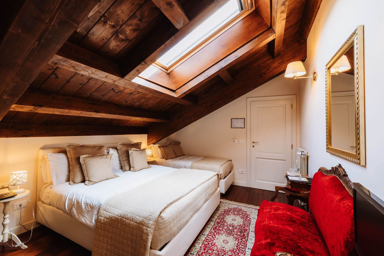 Cozy attic bedroom with exposed wooden beams, skylights, and red accent pillows on white bedding.