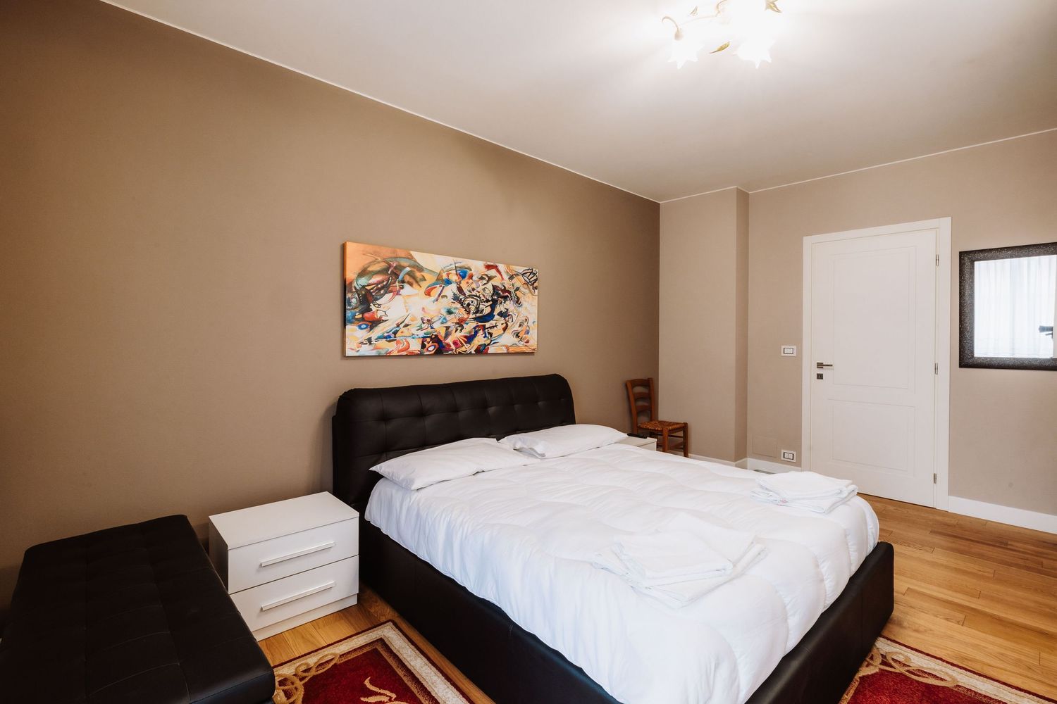 Modern bedroom with white bedding, hardwood floors, and beige walls features artwork and natural lighting.