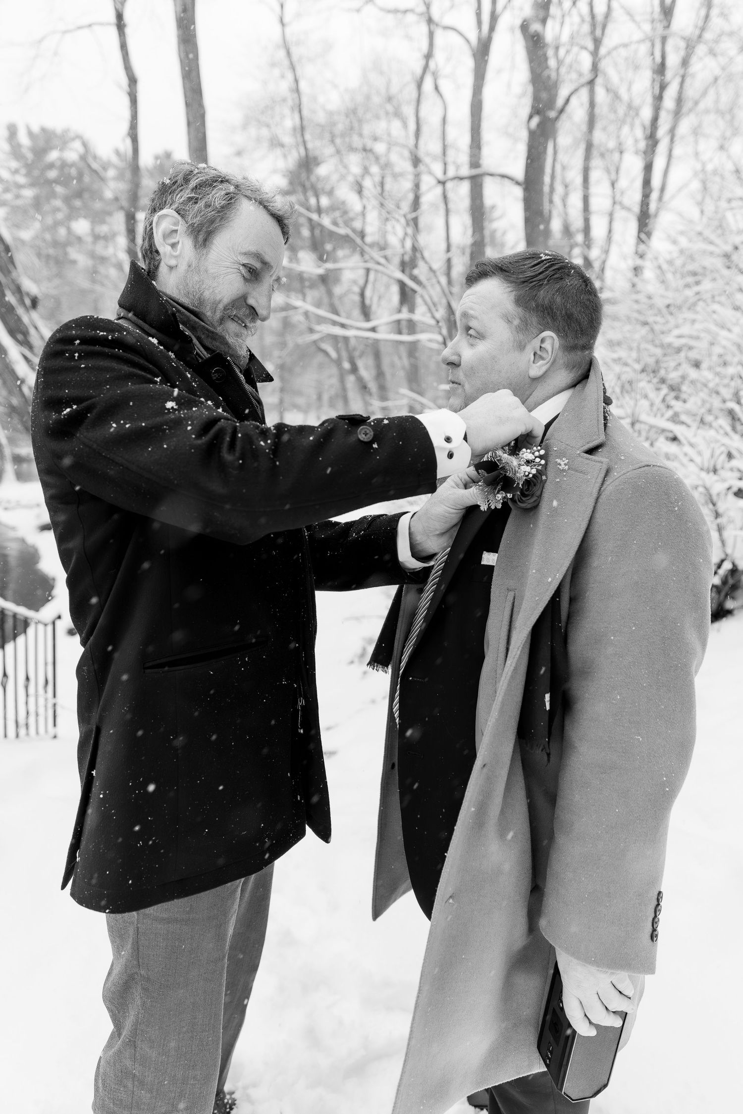 Two men in winter coats, one pinning boutonniere on the other during snowy outdoor wedding.