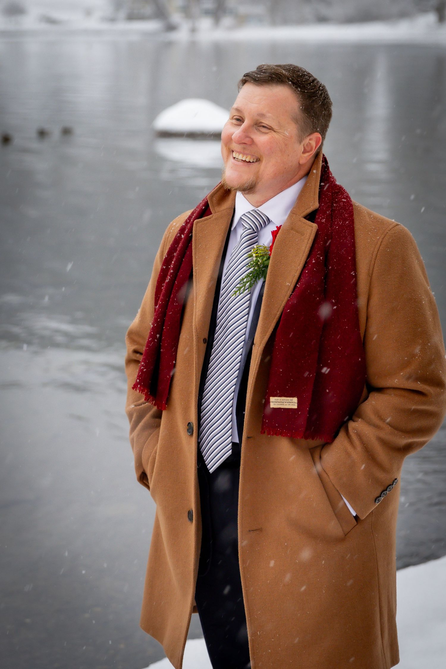 Smiling groom in tan coat and red scarf standing by snowy lake during winter wedding.