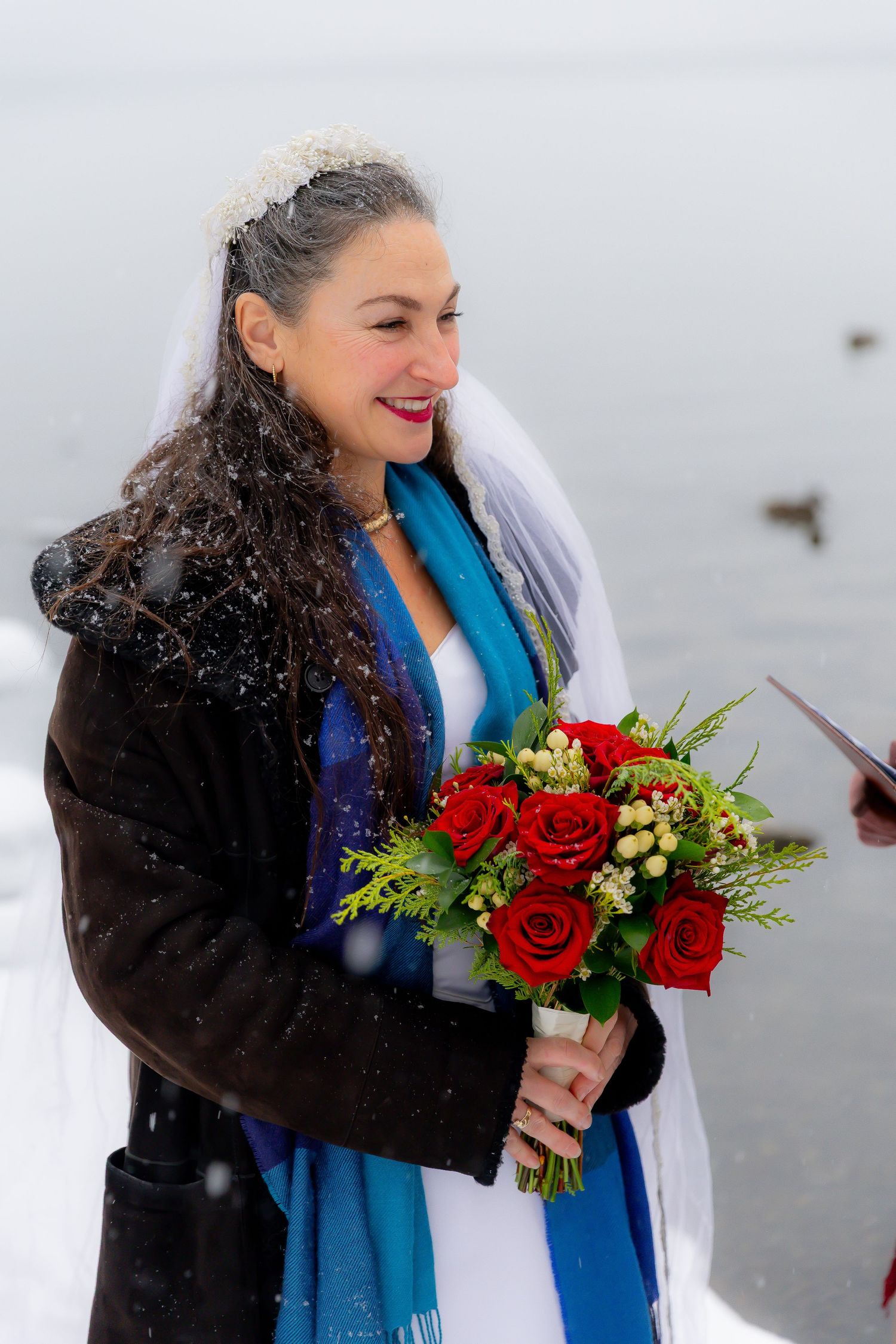 Bride holding red rose bouquet wearing veil and blue scarf during snowy winter wedding.