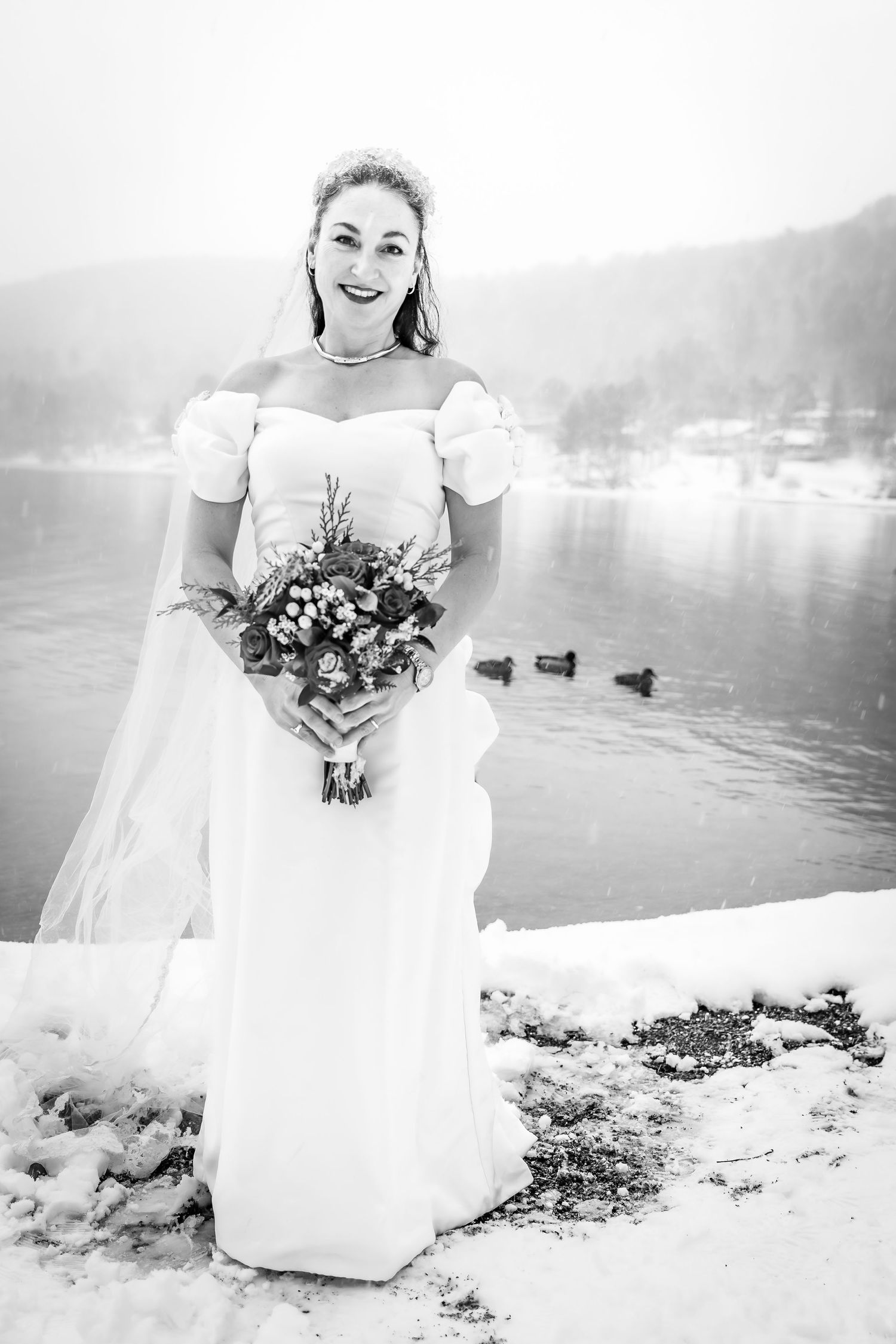Black and white photo of a smiling bride in a white gown holding a bouquet by a snowy lakeside.