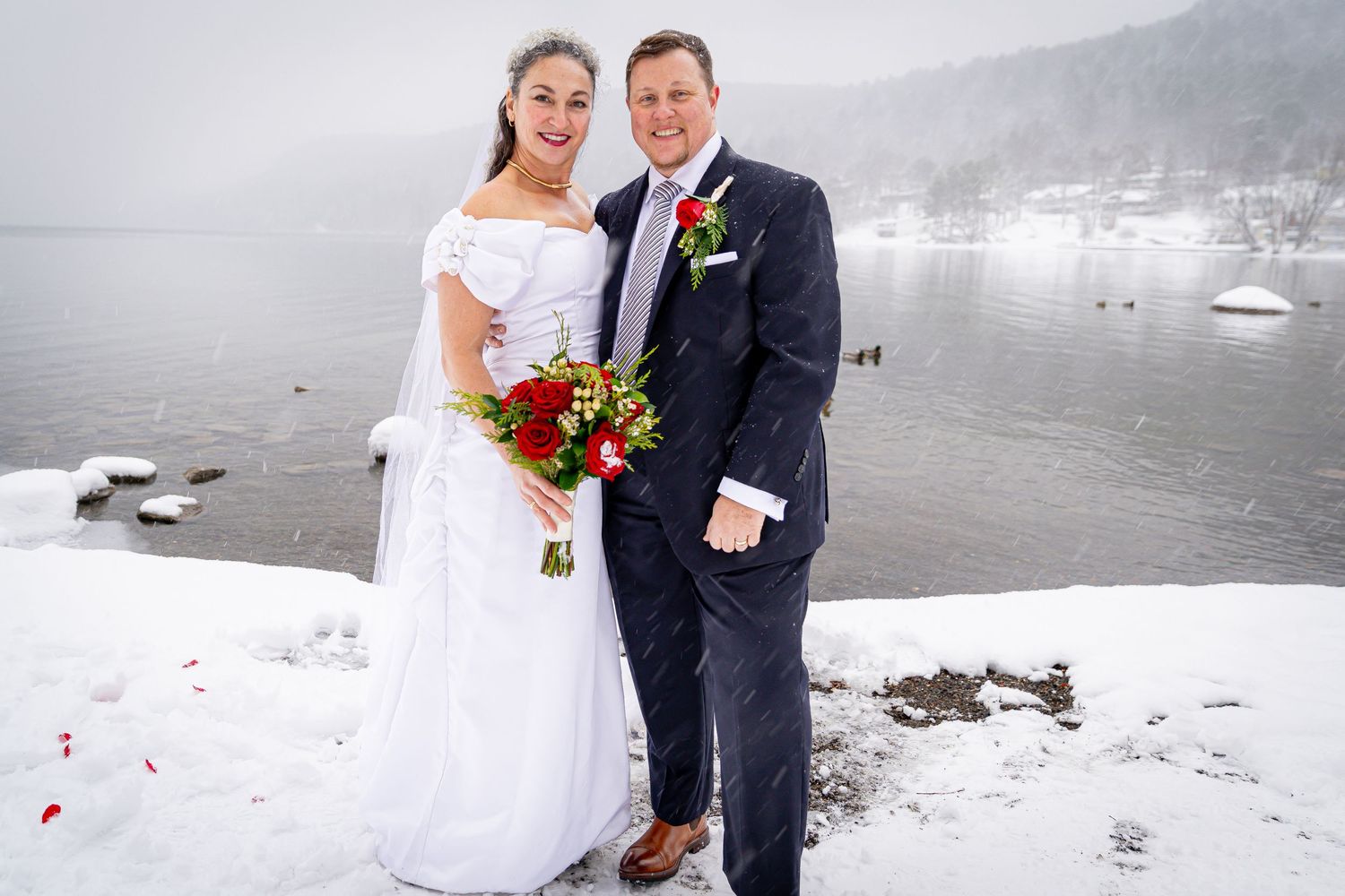 Bride and groom posing together in the snow by a winter lake, bride holding red roses bouquet.