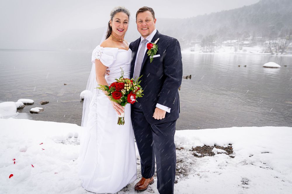 Bride and groom posing together in the snow by a winter lake, bride holding red roses bouquet.
