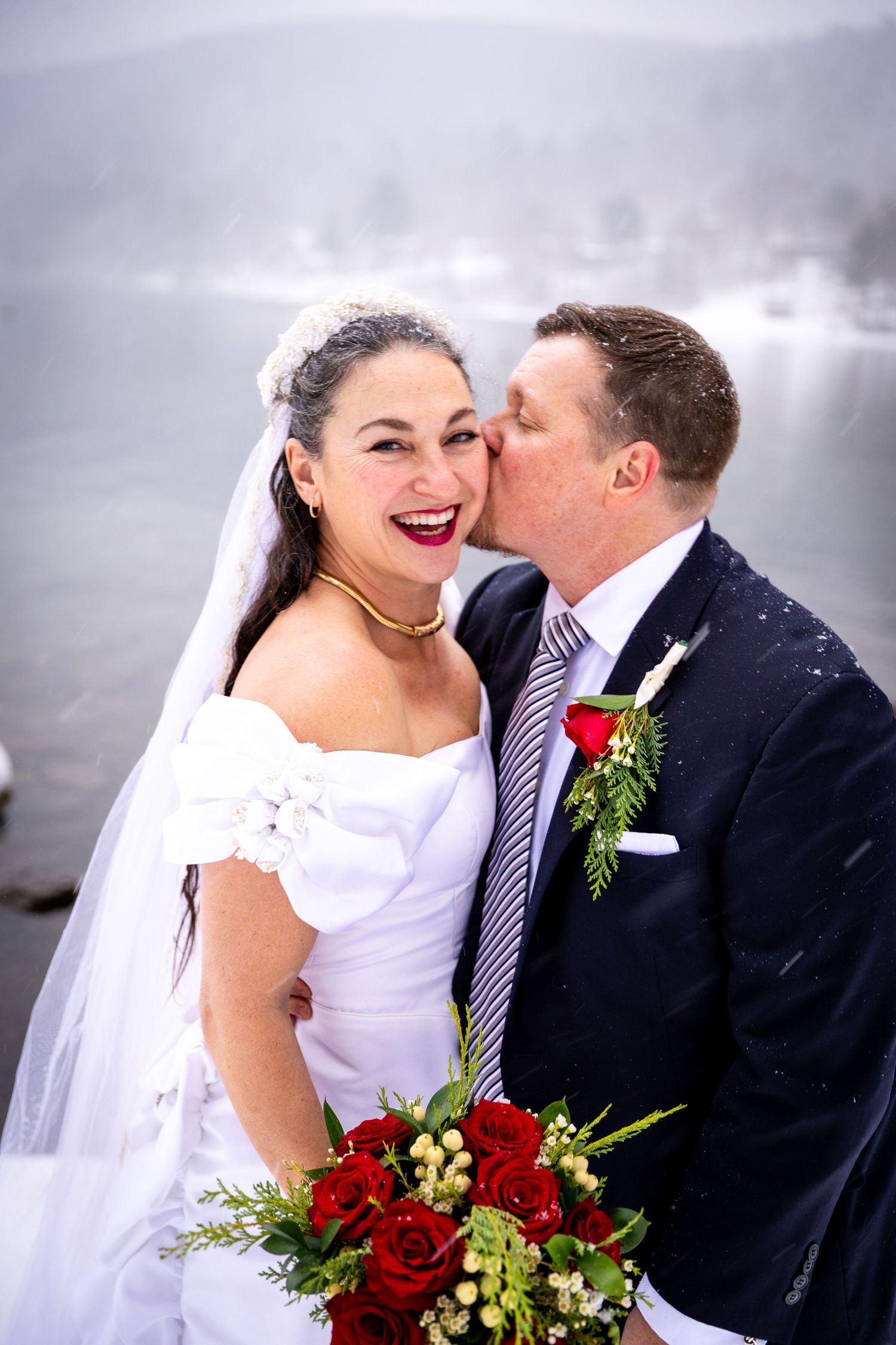 Groom kissing bride on the cheek as she laughs, holding red roses, with a snowy lake backdrop.